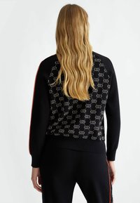 Maglione nero a maglia con schiena decorata da cerchi argentati, strisce laterali rosse a contrasto e polsini aderenti. Texture liscia in tutto il capo.