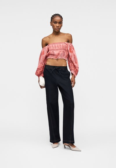 Femme portant un haut court à épaules dénudées rouge à motifs, un pantalon large noir, des talons blancs, et tenant une petite pochette blanche.