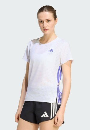 Femme portant un t-shirt de sport blanc perforé avec des rayures violettes et un short noir avec des rayures blanches, debout devant un fond uni.