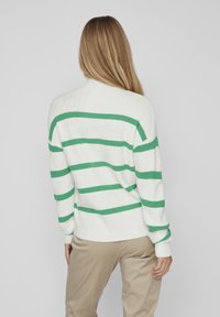 Femme aux longs cheveux blonds portant un pull blanc à rayures horizontales vertes, debout devant un fond uni gris clair.