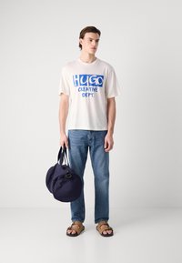 T-skjorte i kremfarge med blå grafisk tekst "HUGO CREATIVE DEPT.", kombinert med blå denimjeans og brune sandaler, holder en marineblå rund skulderveske.