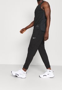Zwarte sportieve tanktop met zij-meshdetails, gecombineerd met zwarte tapered joggers met een wit Nike-logo. Witte sportschoenen met zwarte accenten.