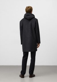 Donkergrijze wollen jas met capuchon, losse pasvorm, knielengte, gestructureerde stof, gecombineerd met donkere broeken en bruine schoenen.