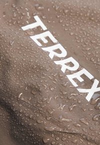 Tissu marron avec le mot « TERREX » imprimé en blanc, recouvert de gouttelettes d’eau dispersées indiquant une résistance à l’eau.