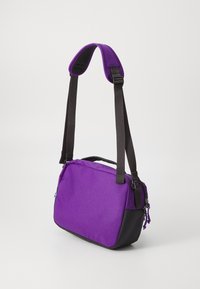 Sac à épaule en tissu violet avec une sangle rembourrée et ajustable. Base noire unie et accents en zip ; forme rectangulaire compacte.