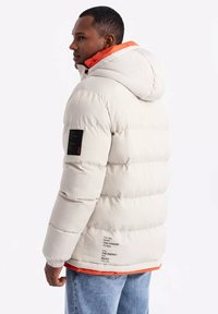 Puffjacka i ljusbeige med en orange innerfoder, som har huva, kviltad textur och en svart etikett på ärmen med tryckt text.