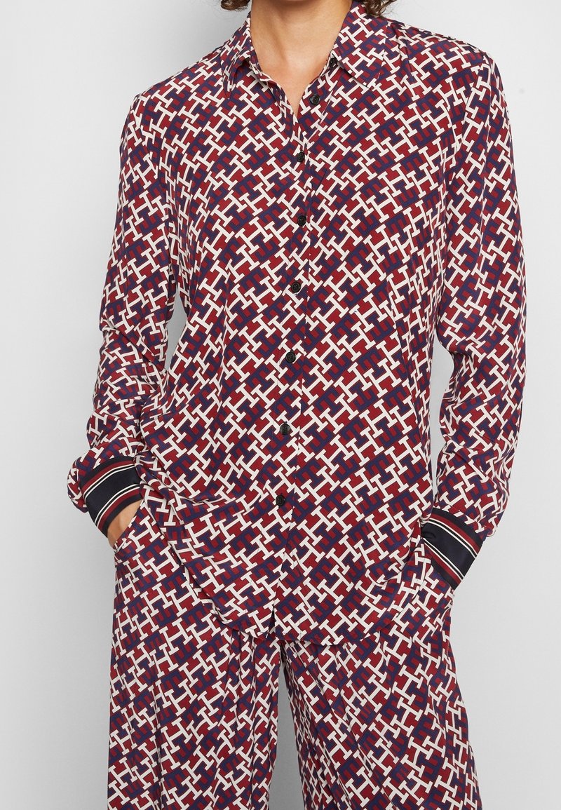 Chemise à manches longues boutonnée et pantalon assorti avec motif géométrique rouge, blanc et marine et poignets rayés noirs, mains du modèle dans les poches.