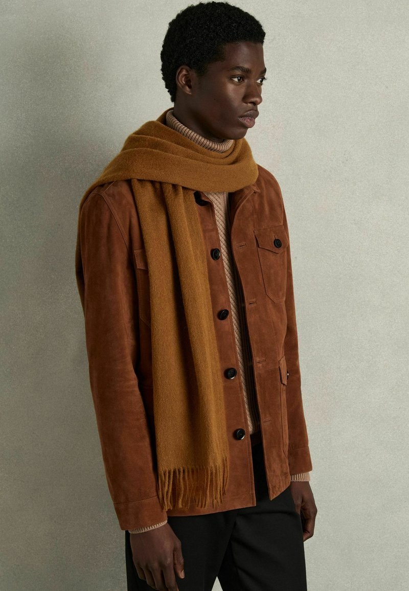 Chaqueta de ante marrón con botones negros, superpuesta a un suéter de cuello alto beige. Combinada con una bufanda mostaza que presenta detalles de flecos.