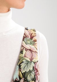 Personne portant un pull à col roulé blanc et une sangle à motif floral sur l'épaule, sur un fond clair et uni.