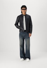 Giacca di pelle nera con colletto alto, tasca con zip frontale, indossata sopra una maglietta in maglia bianca a trama e jeans blu a gamba larga. Sneakers bianche.