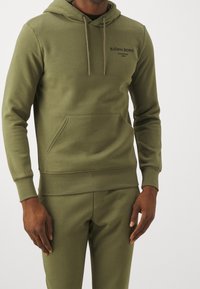 Sudadera color verde oliva con bolsillo frontal, capucha con cordón y mangas ajustadas. Presenta un estampado de logo en la parte superior izquierda.