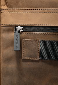 Sezione di borsa in pelle marrone con zip nera, accessori in argento e rifiniture cucite. Include un dettaglio in tessuto texturizzato e un'etichetta con logo.