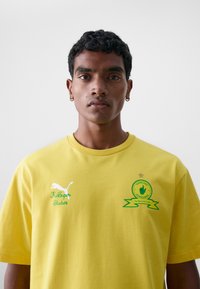 Camiseta de algodón amarilla con cuello redondo, estampados de logotipo en la parte frontal en verde y blanco, y detalles decorativos que incluyen una estrella y cintas.