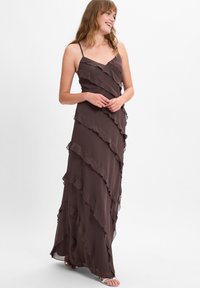 Langes, braunes, ärmelloses Kleid aus fließendem Stoff mit gestuften Rüschen. Das Kleid hat ein V-Ausschnitt-Design und reicht bis zum Boden.