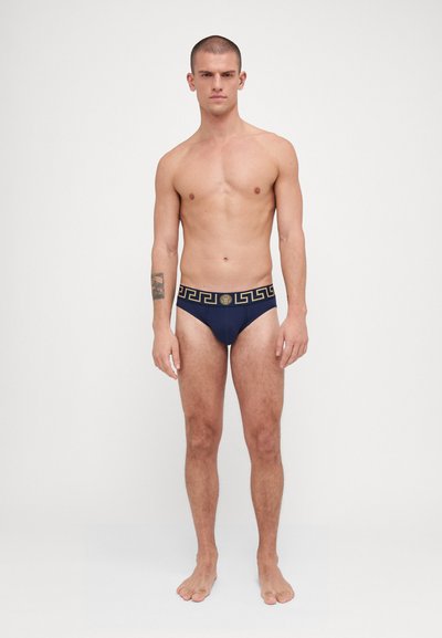 Slip da bagno blu navy con una vita dorata a fantasia con design a chiave greca. Il materiale appare liscio con una forma aderente. Nessun ulteriore dettaglio.