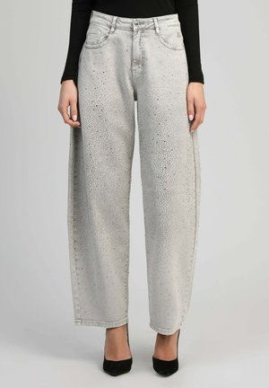 Femme portant un jean gris clair à jambes larges avec un motif à pois subtil, un haut noir et des escarpins noirs à bout pointu, debout devant un fond uni.