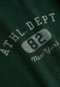 Green ribbed fabric with white text "ATHL. DEPT" and "New York" below a white oval containing the number "82" in dark green.
