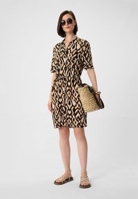 Kurzärmliges, knöpfbares Kleid mit einem tan- und schwarzfarbenen geometrischen Muster, tailliert, kombiniert mit flachen Sandalen und einer geflochtenen Tasche.