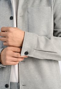 Grå knapp-up jacka med en strukturerad yta, som har en bröstficka, svarta knappar och detaljer med uppvikta ärmar.