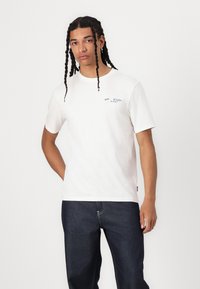 Hvid bomulds t-shirt med korte ærmer, med et blåt logo på venstre bryst og et klassisk crew neck design. Bæret med mørke jeans.