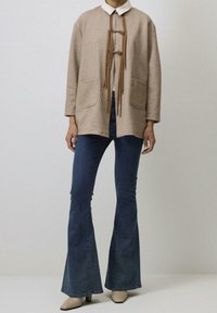 Beżowa kurtka z wełny z kołnierzem, z dwoma przednimi kieszeniami i skórzanym zapięciem na toggle, zestawiona z dzwonowymi dżinsami i beżowymi szpilkami.