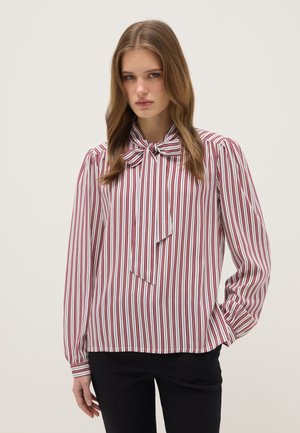 Giovane donna che indossa una camicetta a righe verticali rosse e bianche con un fiocco sul davanti, abbinata a pantaloni neri, in piedi davanti a uno sfondo semplice.