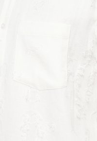 Λευκό πουκάμισο με υφή, διαθέτει τσέπη στο στήθος και διακοσμητικές λεπτομέρειες από ελαφρώς παραγεμισμένο και φθαρμένο ύφασμα.
