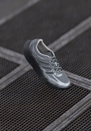 Gray athletic sneaker with thick black sole, laces tied, resting on dark metal grid flooring.
