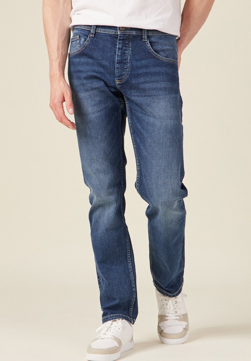 Dunkelblaue Jeans im klassischen Schnitt. Verfügt über fünf Taschen mit Steppnähten und dezenten Verwaschungen. Kombiniert mit weißen Sneakers.