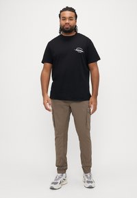 Μαύρο T-shirt με στρογγυλή λαιμόκοψη και κοντά μανίκια, με μικρό λευκό σχέδιο στο αριστερό στήθος· συνδυασμένο με χακί παντελόνι cargo και γκρι αθλητικά παπούτσια.