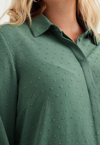 Grön skjorta med krage i strukturerat tyg som har upphöjda prickar. Lätt material, knappstängning och avslappnad passform.