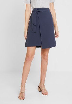 Falda cruzada azul marino hecha de un material suave, con un lazo lateral y un dobladillo recto. Se lleva con una blusa gris y tacones beige con tiras.