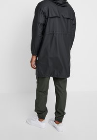 Czarny wodoodporny płaszcz z kapturem, elastycznymi mankietami i rozcięciem z tyłu, zestawiony z zielonymi spodniami cargo i białymi sneakersami. Prosty, funkcjonalny design.