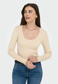 Beige ribbeltrui met lange mouwen en een scalloped halslijn, gecombineerd met blauwe jeans. De stof heeft een gladde textuur en een aansluitend ontwerp.