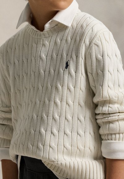 Enfant portant un pull crème à torsades par-dessus une chemise blanche à col, avec les manches remontées et un pantalon foncé visible en dessous.