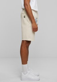 Shorts beige clair en tissu doux avec une taille élastique, des poches latérales et des détails graphiques bleus, associés à des baskets blanches et des chaussettes.