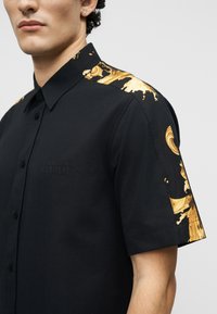 Νεαρός άνδρας που φοράει μαύρο πουκάμισο με κοντά μανίκια και χρυσά μοτίβα μπαρόκ στο στυλ στους ώμους και τα μανίκια, στέκεται μπροστά από ένα απλό φόντο.