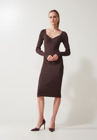 Ribbed, brown long-sleeve dress with a fitted silhouette and a v-neckline, paired with glossy burgundy stiletto heels.