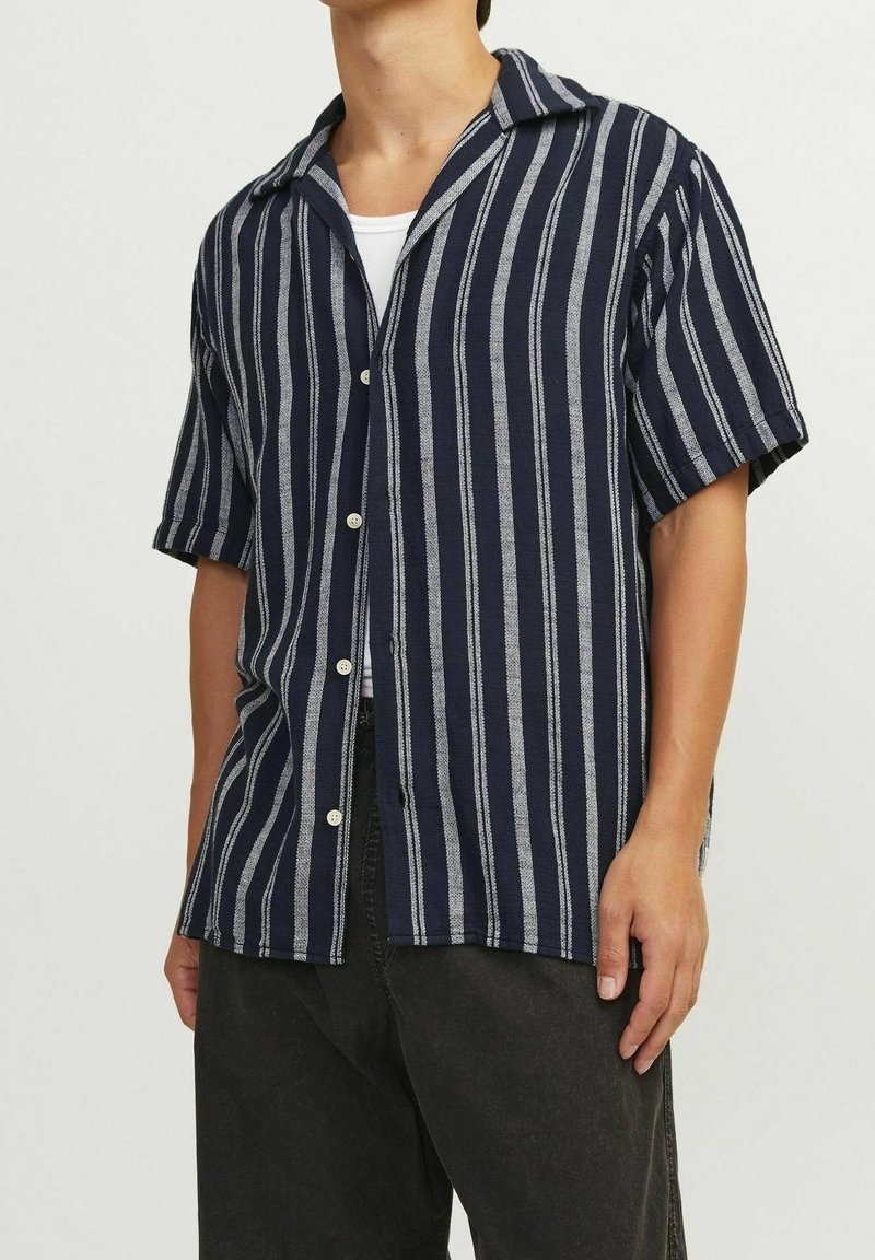 Granatowo-biała, w paski, koszula z krótkim rękawem, z kołnierzykiem, zapinana na guziki i w strukturze materiału. Nosi się ją na białej bluzce na ramiączkach.