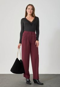 Zwart top met lange mouwen en kanten halslijn, gecombineerd met hoge taille bordeauxrode broek en een zwarte suède tas. Het model draagt zwarte puntige enkellaarzen.