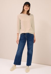 Frau trägt ein beiges Langarmoberteil, dunkelblaue, weite Jeans mit Knöpfen, beige Sneakers und eine schwarze Baseballmütze. Sie steht vor einer schlichten Wand.