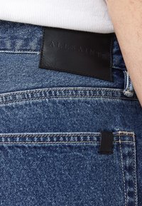 Farkut denim sinisessä pesussa, joissa on musta nahkaleima, johon on kohokuvioitu "ALLSAINTS", ja erottuvat tikkaus takataskuissa.