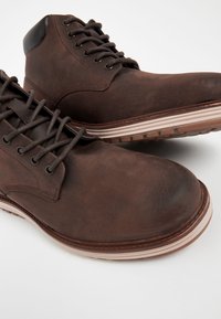 Hnedé kožené členkové topánky s robustným dizajnom, okrúhlym špicom, predným zaväzovaním na šnúrky a svetlou gumovou podrážkou s textúrovaným dezénom.