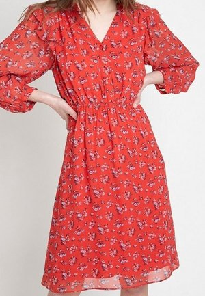 Frau trägt ein knielanges rotes Blumenmusterkleid mit langen Ärmeln und geschnürter Taille, steht mit den Händen in die Hüften gestützt.