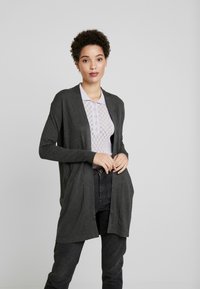 Dunkelgrauer Cardigan mit offener Vorderseite, langen Ärmeln und lässiger Passform. Trägt man über einem helllila Kragenoberteil mit strukturiertem Muster.
