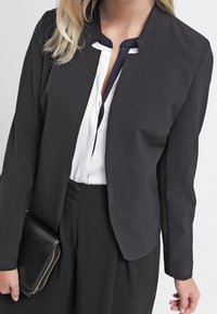 Veste noire structurée à manches longues, sans revers, avec une silhouette ajustée, portée sur une chemise blanche avec un col marine.