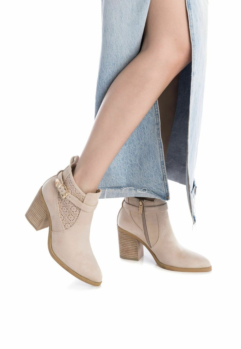 Botas de tobillo beige con detalles perforados, puntera redonda y tacón apilado. Cierre de cremallera y acento de hebilla. Material de ante texturizado.