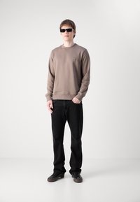 Sudadera marrón con cuello redondo y mangas acampanadas, combinada con jeans negros y zapatillas negras. Las gafas de sol completan el atuendo.
