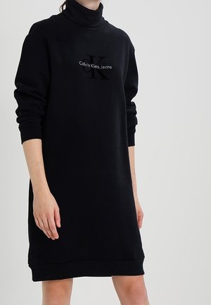 Robe noire à manches longues avec col roulé, poignets retombés et logo Calvin Klein Jeans sur la poitrine, portée par une personne debout sur un fond uni.