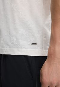 Witte shirt met korte mouwen met een textuurpatroon van geperste diamanten en bloemvormen, voorzien van een zilverkleurig logo-label nabij de zoom.